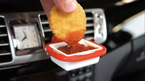 An image of a cup clipped to a car vent. There is a sauce packet inside the cup and someone is dipping a nugget into it. The product is called the Saucemoto.