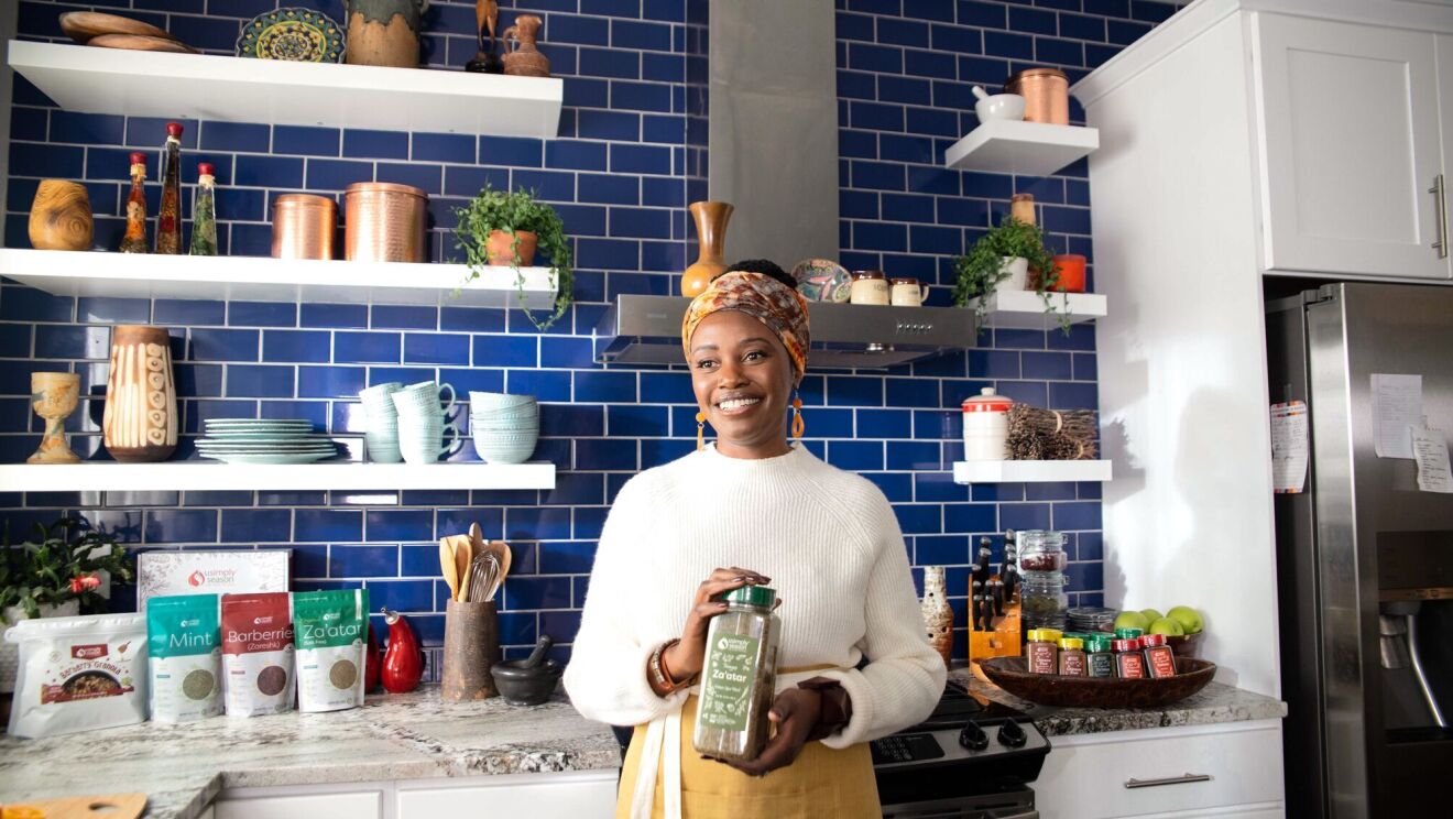 sylvia kapsandoy the founder of usimplyseason stands in front of a kitchen stove and countertop holding a carton of spice seasonings 