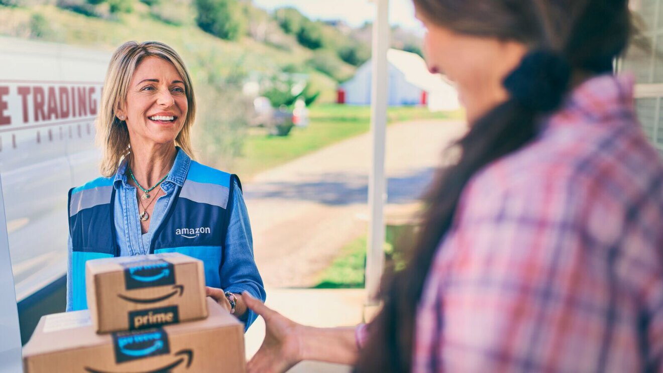 An image of an Amazon delivery driver handing two prime boxes to a customer standing at her doorstep.