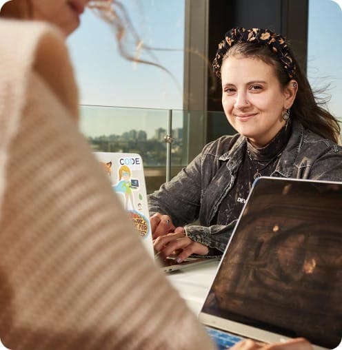 Mujer sonriendo mientras mira una computadora frente a otra persona que no se llega a ver completa, también en una computadora