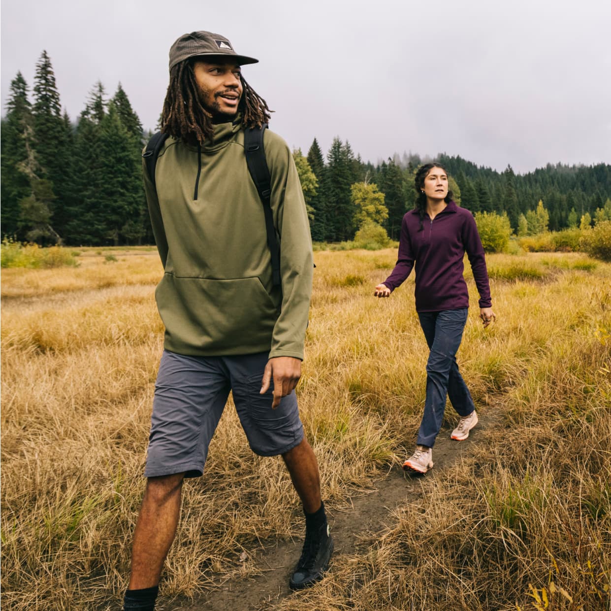 Hikers walk through a grassy field bordered by pine forest.