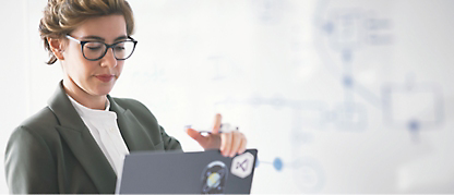 A woman in glasses is holding a laptop in front of a whiteboard.