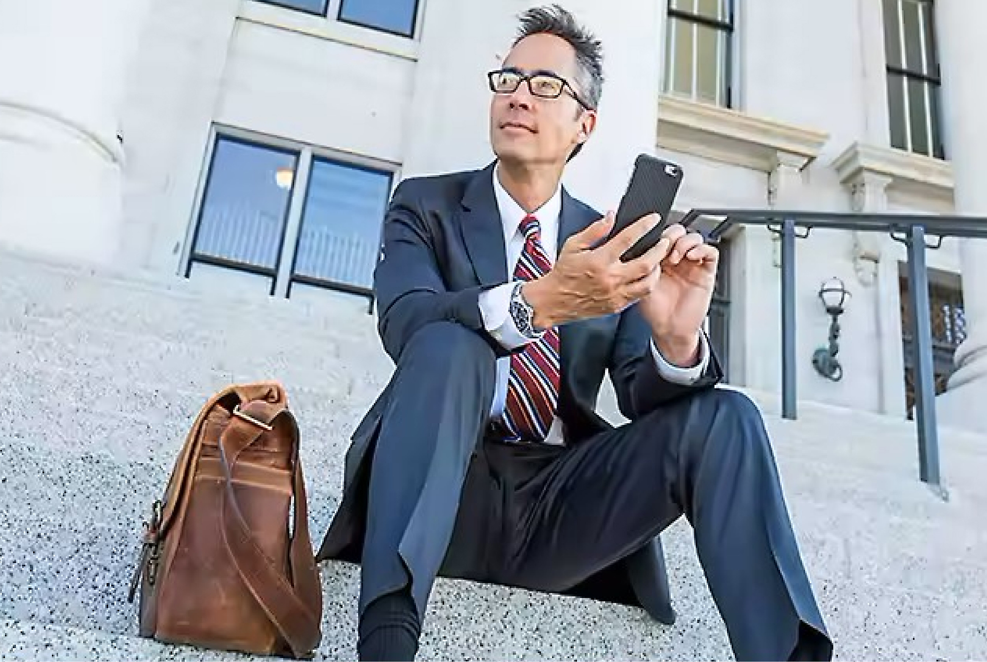 スーツを着て眼鏡をかけたビジネスマンが、建物の外の階段に座り、革製の鞄を横に置いてスマートフォンを使っています。