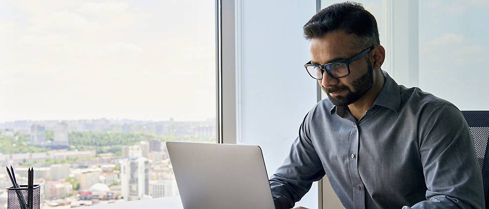 Muž s brýlemi, který pracuje na notebooku v kanceláři ve výškové budově s výhledem na město v pozadí