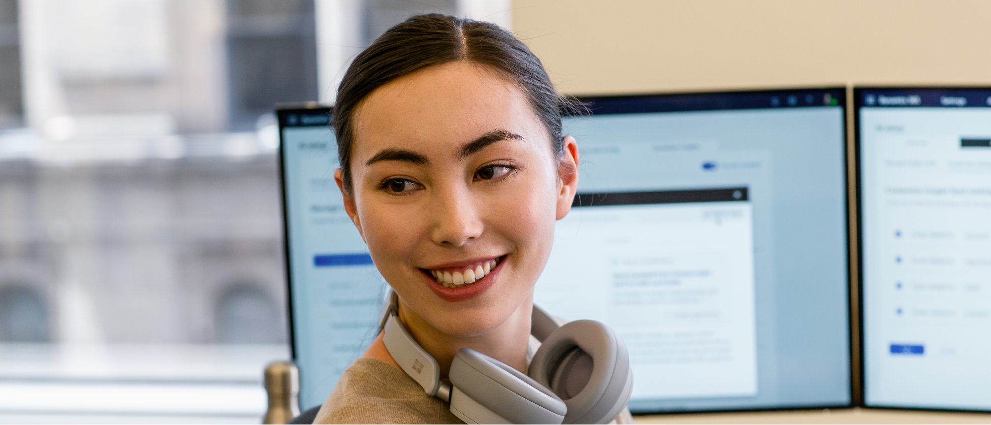 Μια γυναίκα τοποθετεί τα ακουστικά της πάνω από τον λαιμό της και χαμογελώντας ενώ εργάζεται σε δύο οθόνες.