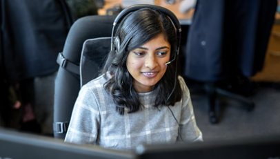 Een vrouw die aan een bureau achter een beeldscherm zit en een headset draagt.