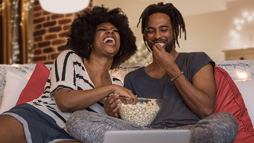 To personer som ler og spiser popcorn mens de ser på TV.