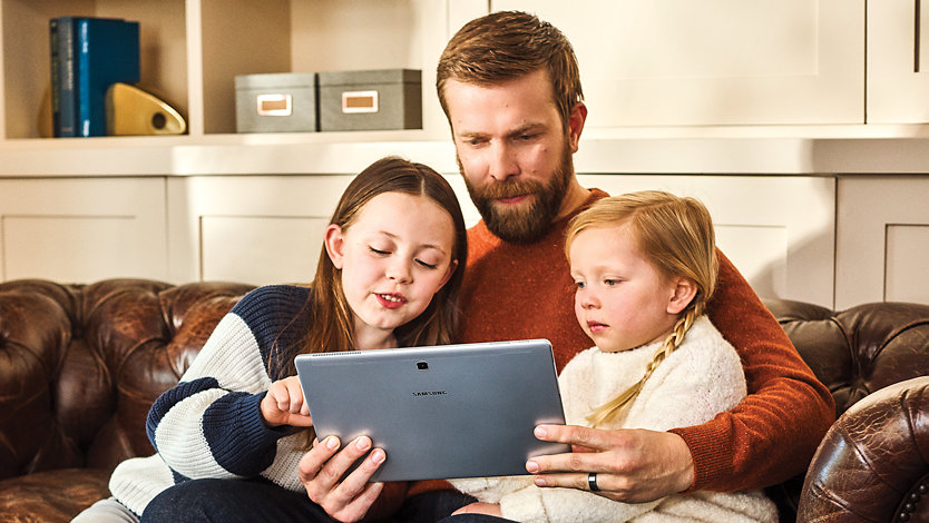 A bearded man holds his daughters close as they look at something on a tablet.