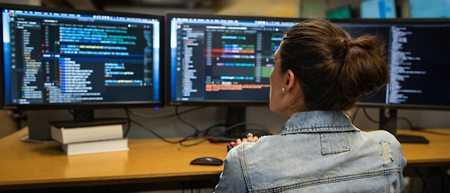 Žena, která v tmavé kanceláři programuje na několika monitorech, na kterých se zobrazuje kód