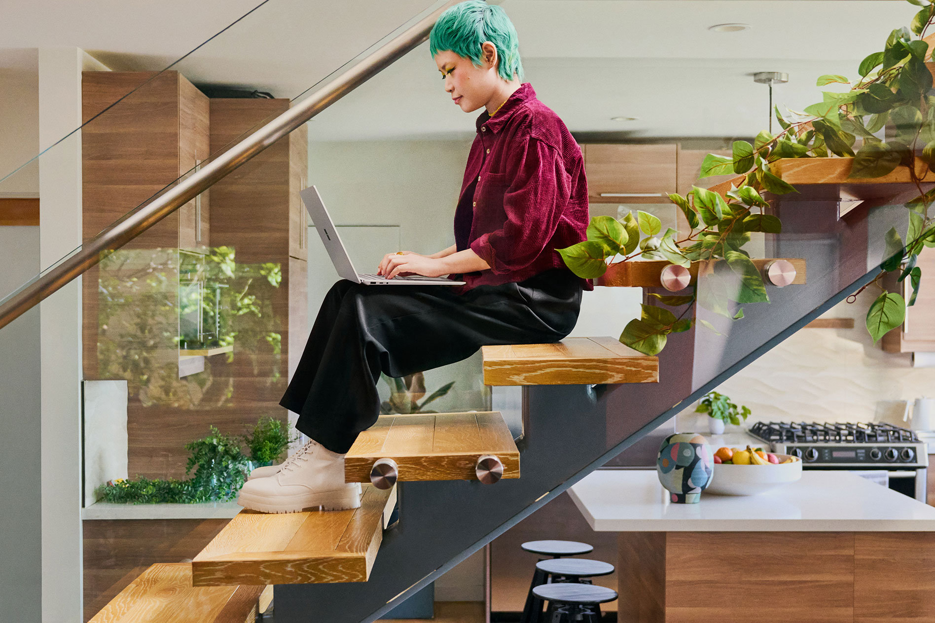 Une jeune personne assise sur un escalier dans une cuisine moderne tape sur le clavier de son Surface Laptop 5.