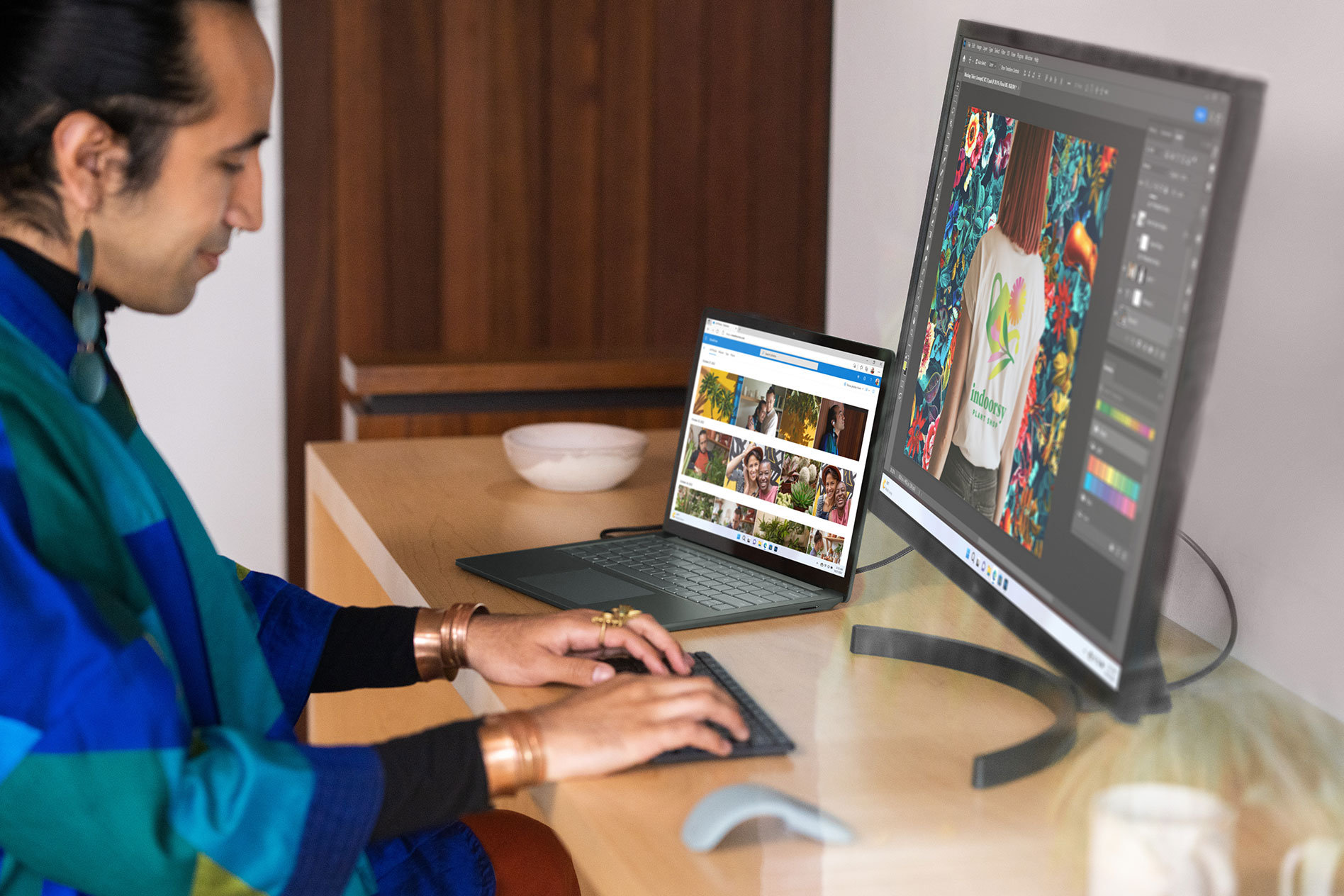Een persoon zit aan een houten bureau te typen op een extern toetsenbord met zowel de Surface Laptop 5 als een externe monitor die foto's op het scherm toont.