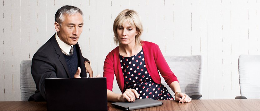 Ένας άντρας και μια γυναίκα κάθονται σε ένα τραπέζι συσκέψεων και κοιτάζουν έναν φορητό υπολογιστή.
