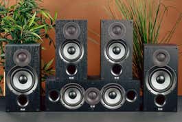 The Elac Debut 2.0 speaker system, shown arranged against a brown background with houseplants behind the speakers.