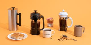 An array of different french press coffee makers, together with coffee mugs, coffee beans, jam, and toast.