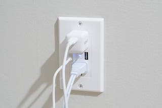 The Geeni Current+Charge in-wall smart outlet, shown installed in a wall, with two devices plugged into the sockets and a USB charging cable in one of the two USB ports.