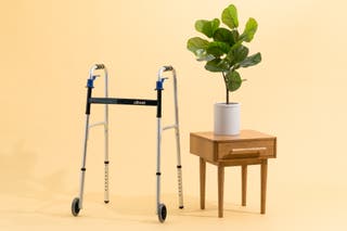 The Drive Deluxe Trigger Release Folding Walker, next to a potted plant in front of a beige background.