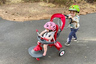A kid pushing another kid on a tricycle.