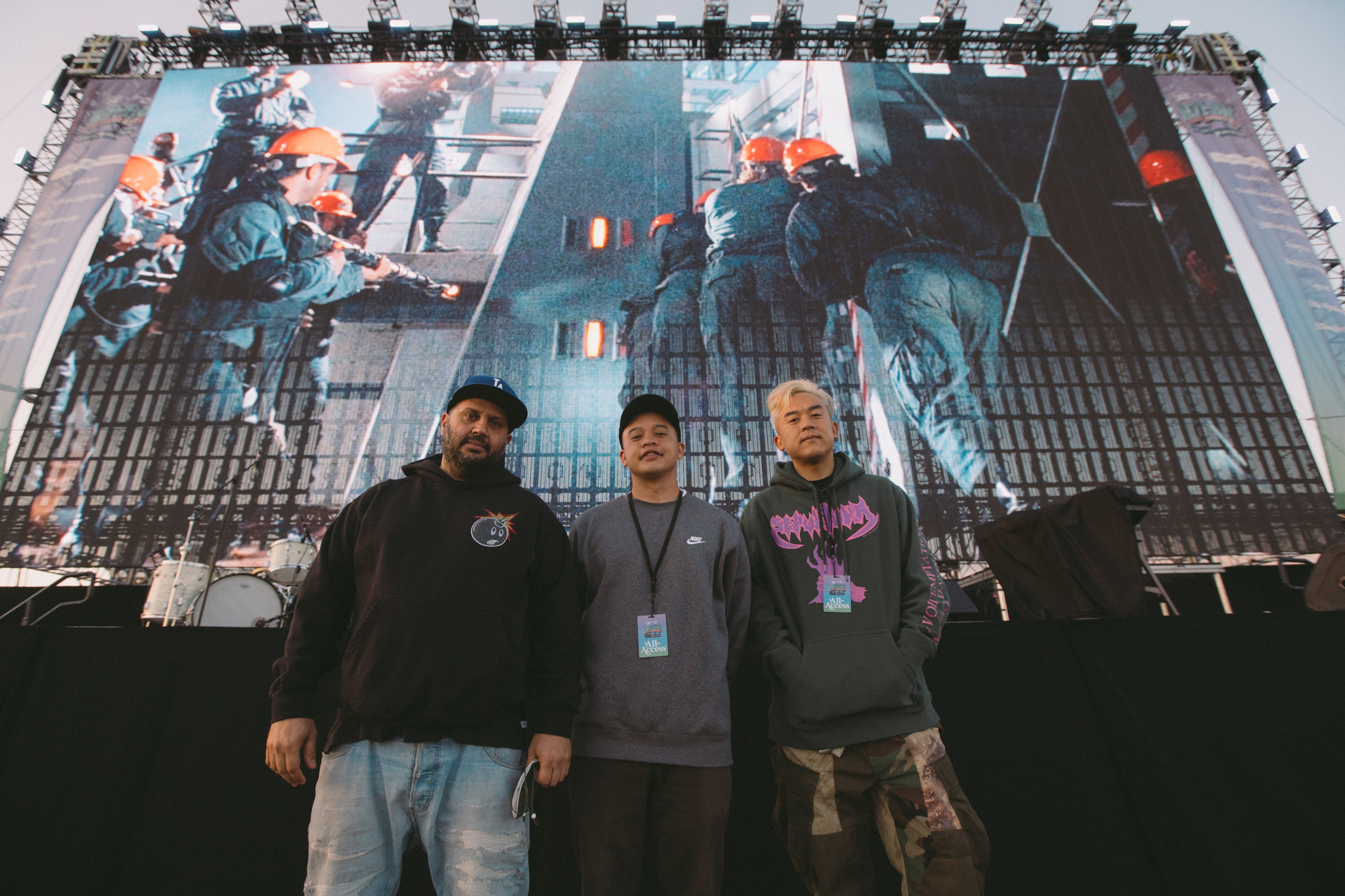 Three men stand wearing streetwear in front of a music performance stange.