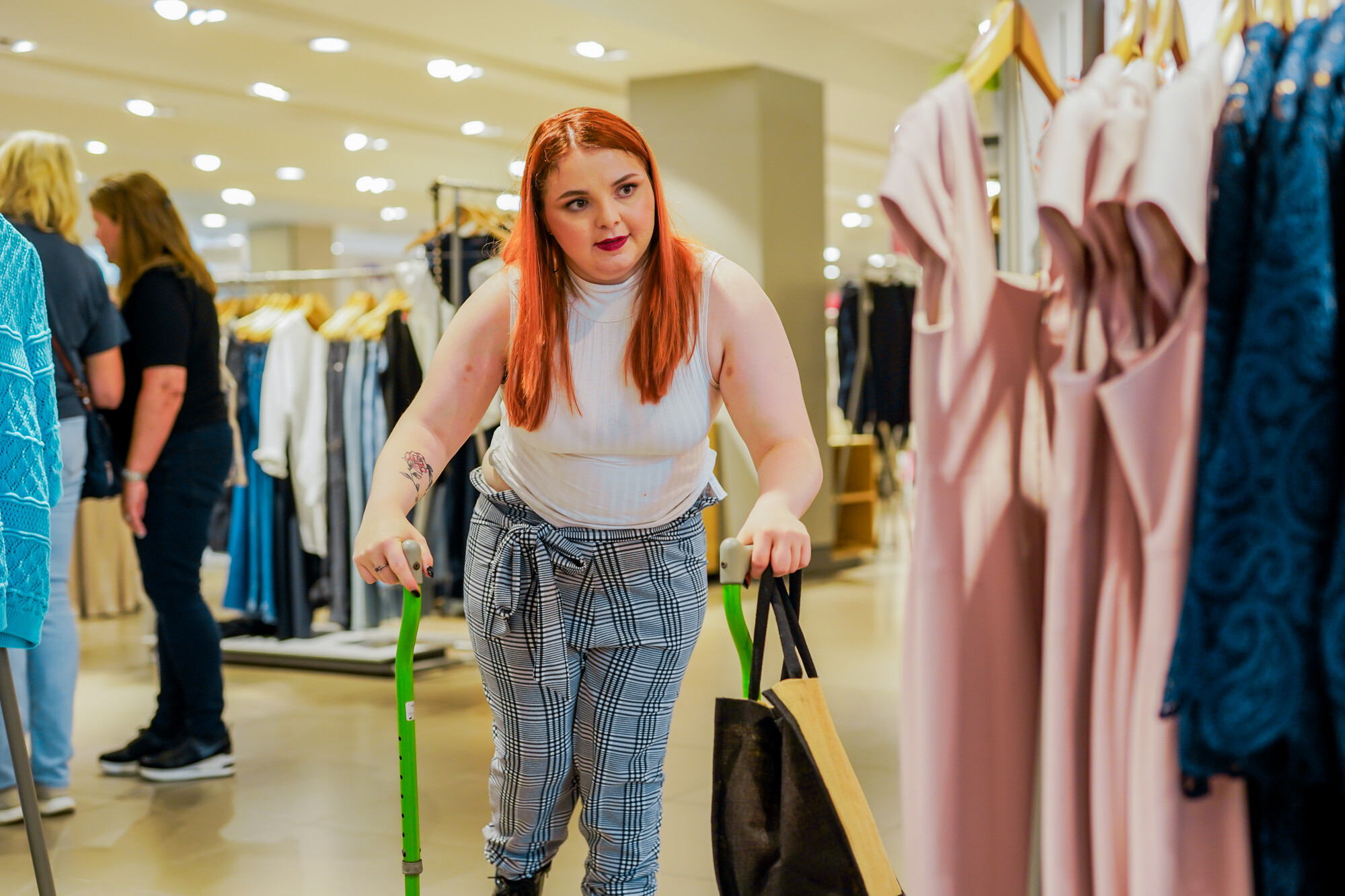 Woman with long red hair and bright green walking sticks looks at dresses in a clothes store. She has a tattoo of a rose on her right arm.