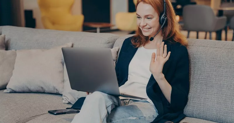 Red haired woman sitting on sofa greeting an online meeting.