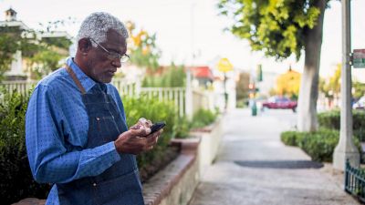 Hombre mayor usando un delantal de artesano, enviando un mensaje de texto en su teléfono inteligente en una calle de barrio