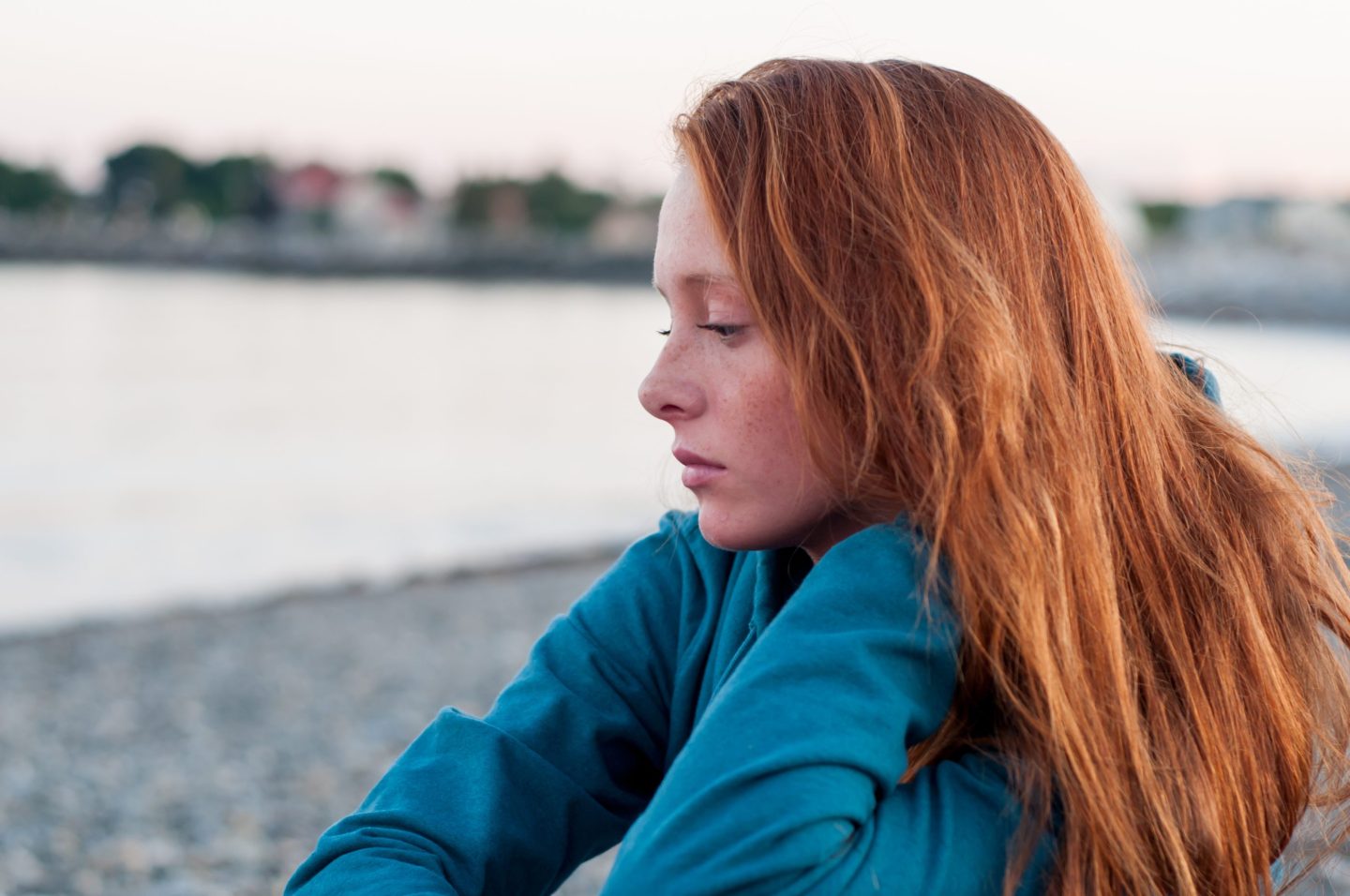 Redheaded teenager in a blue shirt sitting at the ocean and looking contemplative.