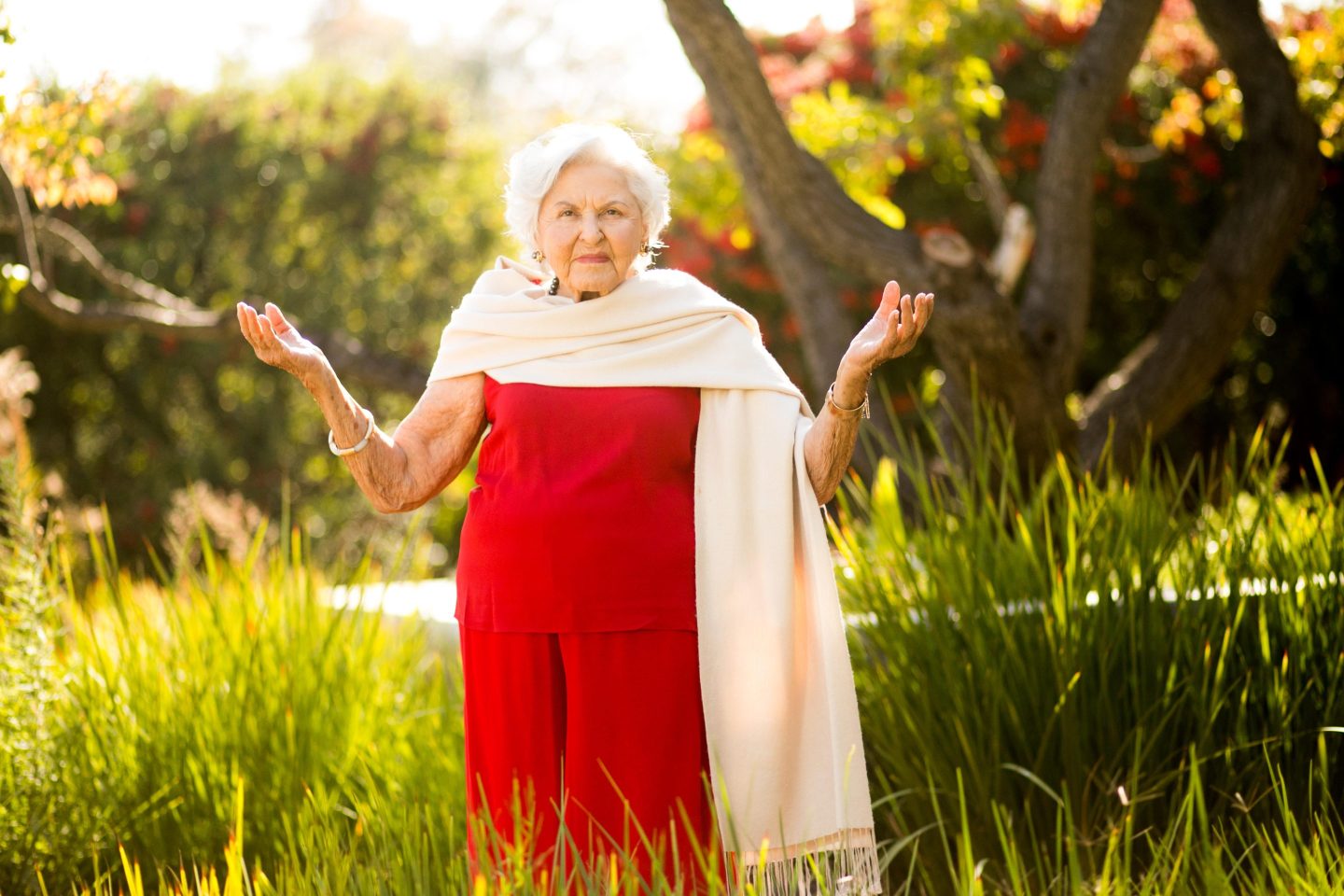An elderly woman in a red dress standing in tall grass with her arms outstretched