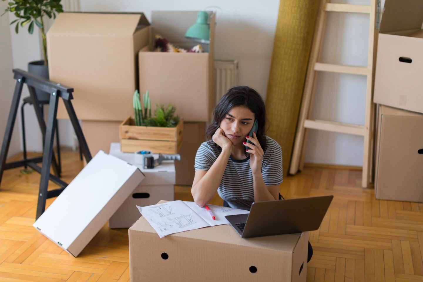 Beautiful young Caucasian woman moving into her new home and starting a new life, taking a break with the unpacking to finish some business first, using a laptop and a smart phone, feeling tiered and bored