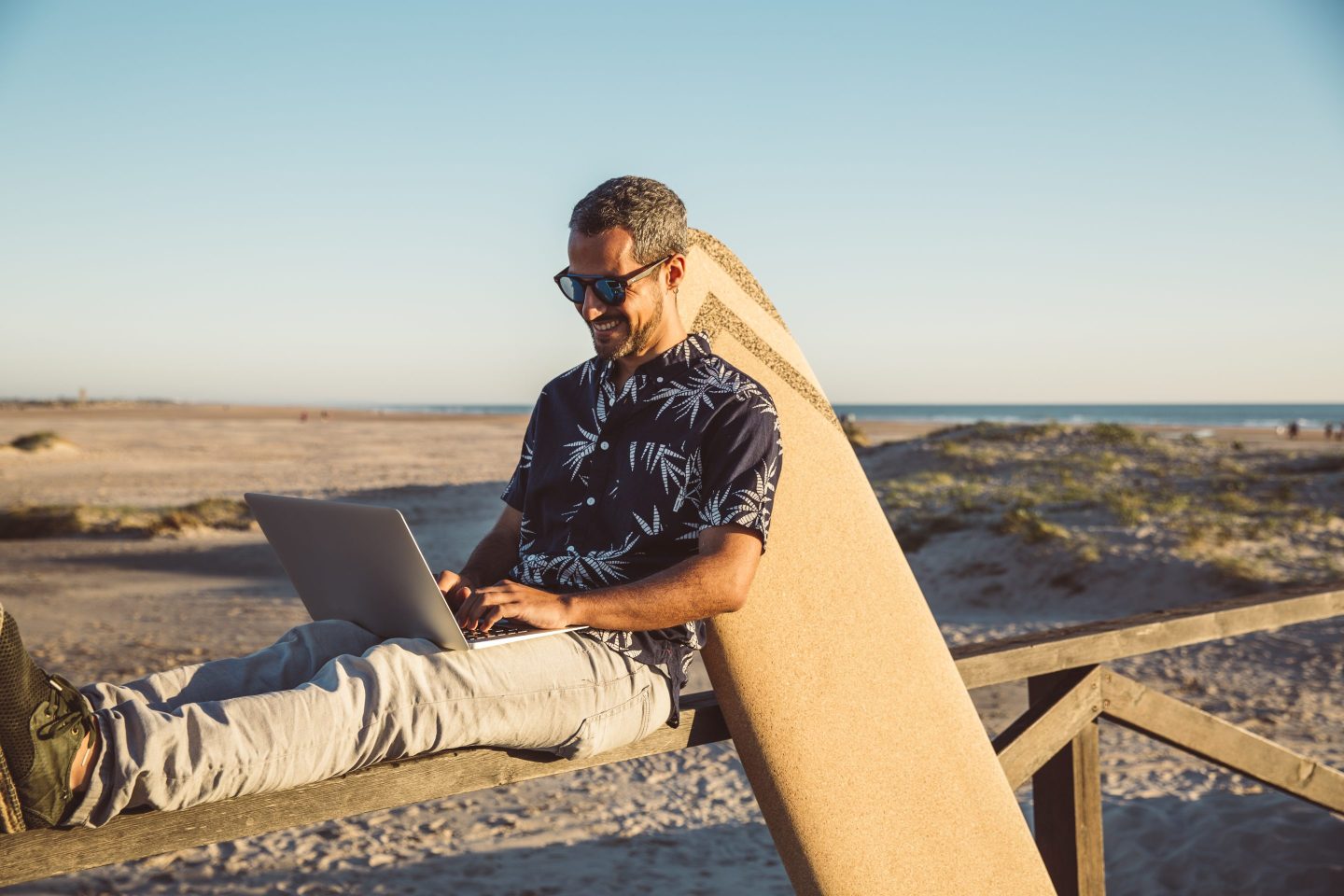 Man sitting at the beach, using laptop, with surfboard leaning on fence