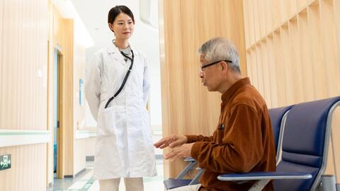 Photo of a doctor and patient in a hospital setting. The doctor wears a white coat and has a stethoscope hanging around her neck. The patient sits on a blue chair and wears a brown shirt. The background is a wooden wall. This photo is taken from the patient's perspective. The location is in China.