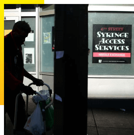 photo of a man with a walker outside of a building with a sign that reads, "6th Street Syringe Access Services, Needle Exchange."