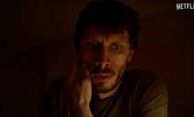 A close-up of a man's face as he stares down at a computer in a dark room.