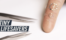 A white background photograph shows a couple of medical tweezers next to three of the tiny stickers placed on an index finger for scale.