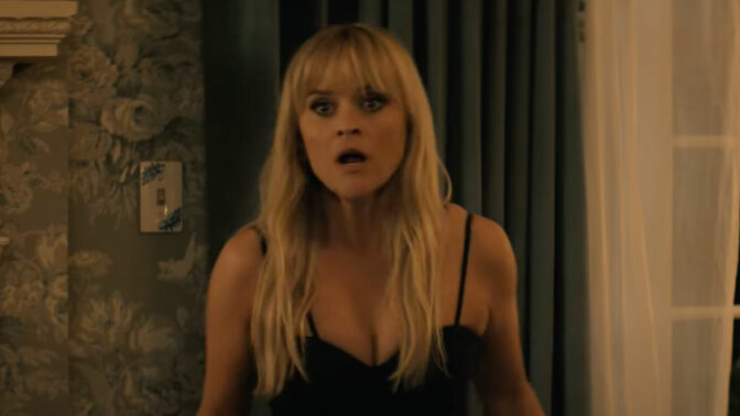 A woman stands in a bedroom with her mouth open, looking shocked.