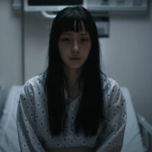 A woman sits in a hospital bed wearing a hospital gown, staring straight at the camera.