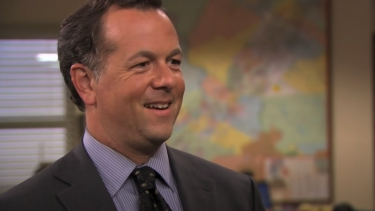 A man (David Costabile as the banker on "The Office") standing in an office wearing a suit and smiling.