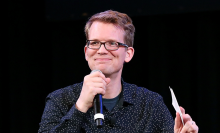 Hank Green in a blue button up shirt, holding a microphone.