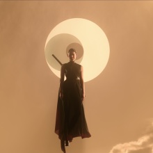 A woman with a sword at her back floats through an orange sky against a symbol of three interlocking circles.