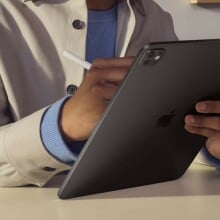 a person holds the apple ipad pro in their left hand while writing with an included e-pen with the right hand. the person is wearing a khaki colored jacket with a blue sweater underneath.