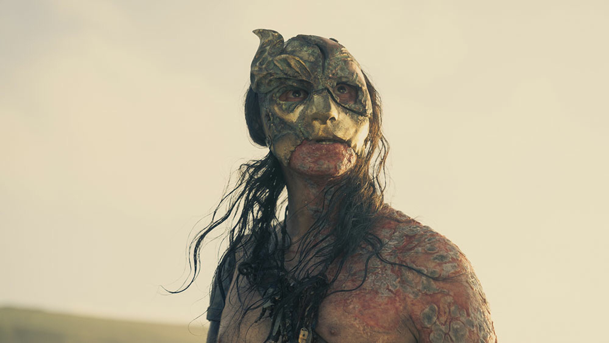 A man with long hair stands shirtless outdoors, wearing a mask.