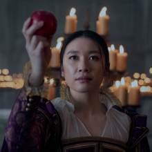 A woman in a 17th century England dress holds out an apple.