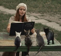 a woman sitting at a table reading a book with cats around her