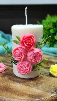 a white candle with pink roses on it sitting on a wooden table next to flowers