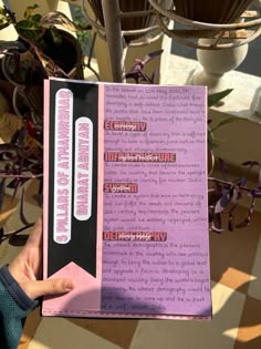 a person holding up a pink book in front of a potted plant with purple flowers