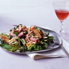 a salad with meat and greens on a plate next to a glass of wine