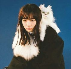 a woman holding a white cat on her shoulders and wearing a black coat over her shoulder
