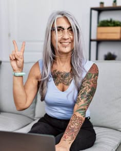 a woman with grey hair and tattoos sitting on a couch holding up her hand in the air