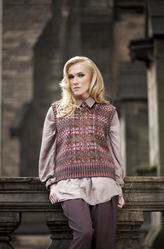 a woman standing next to a stone wall wearing a sweater and pants with buttons on the side