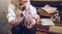 a woman with tattoos on her arms and legs sitting in front of a desk full of papers
