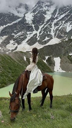 a woman riding on the back of a brown horse next to a lake and snow covered mountains